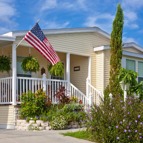 The front of a mobile home surrounded by foliage with an American flag outside.